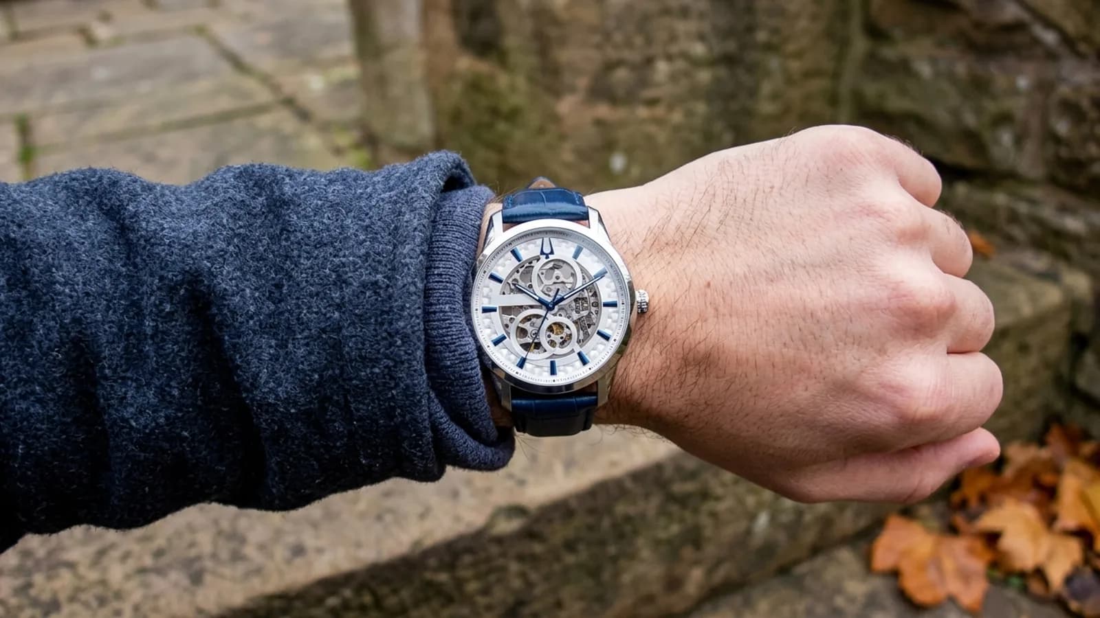 Skeleton-dial stainless steel watch on a blue leather strap, worn over a navy sweater cuff on an autumn stone step.